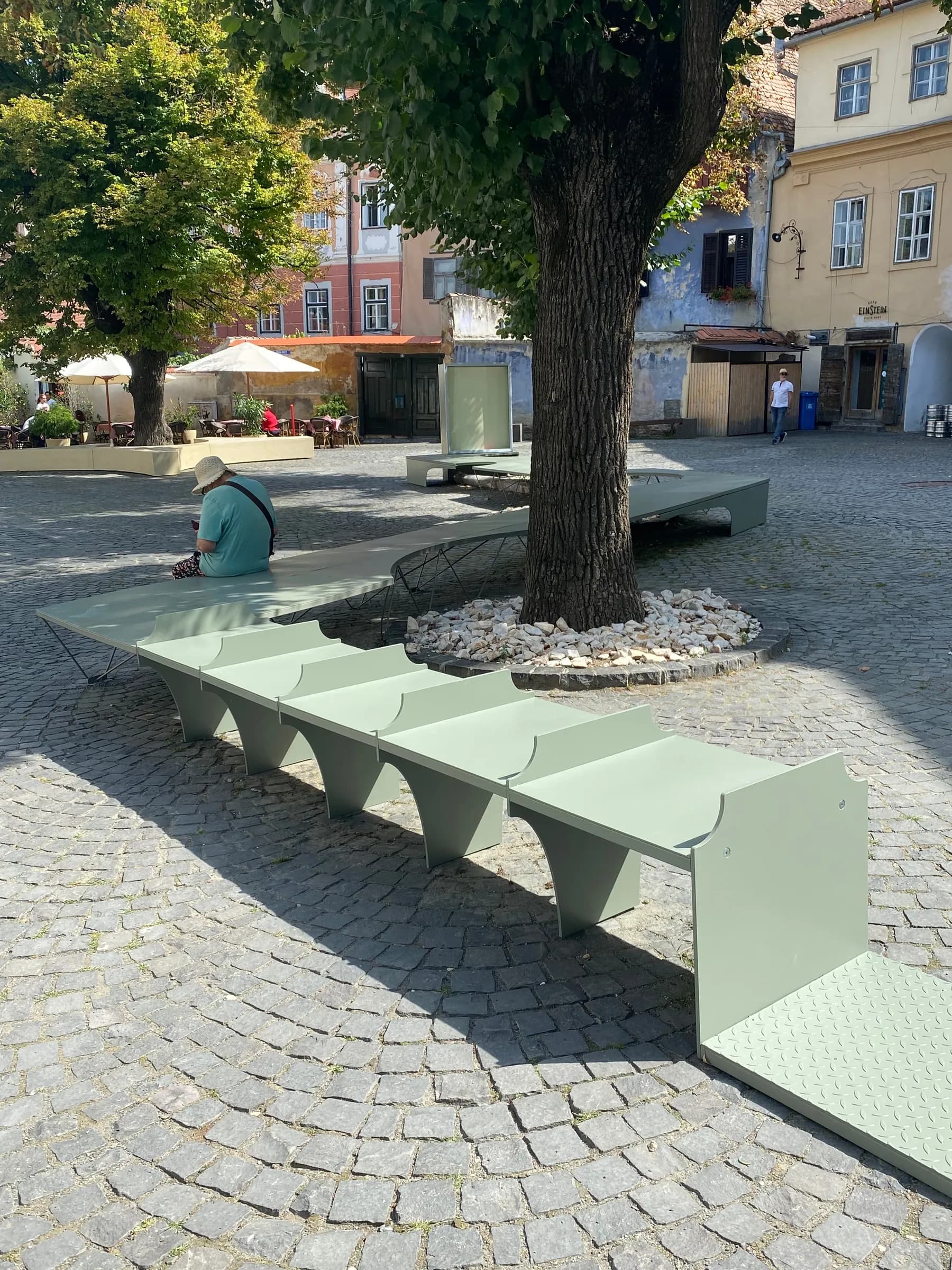 Greenish-grey steel bench of seat modules with curved edges, continuing into a low platform on rod legs around a tree