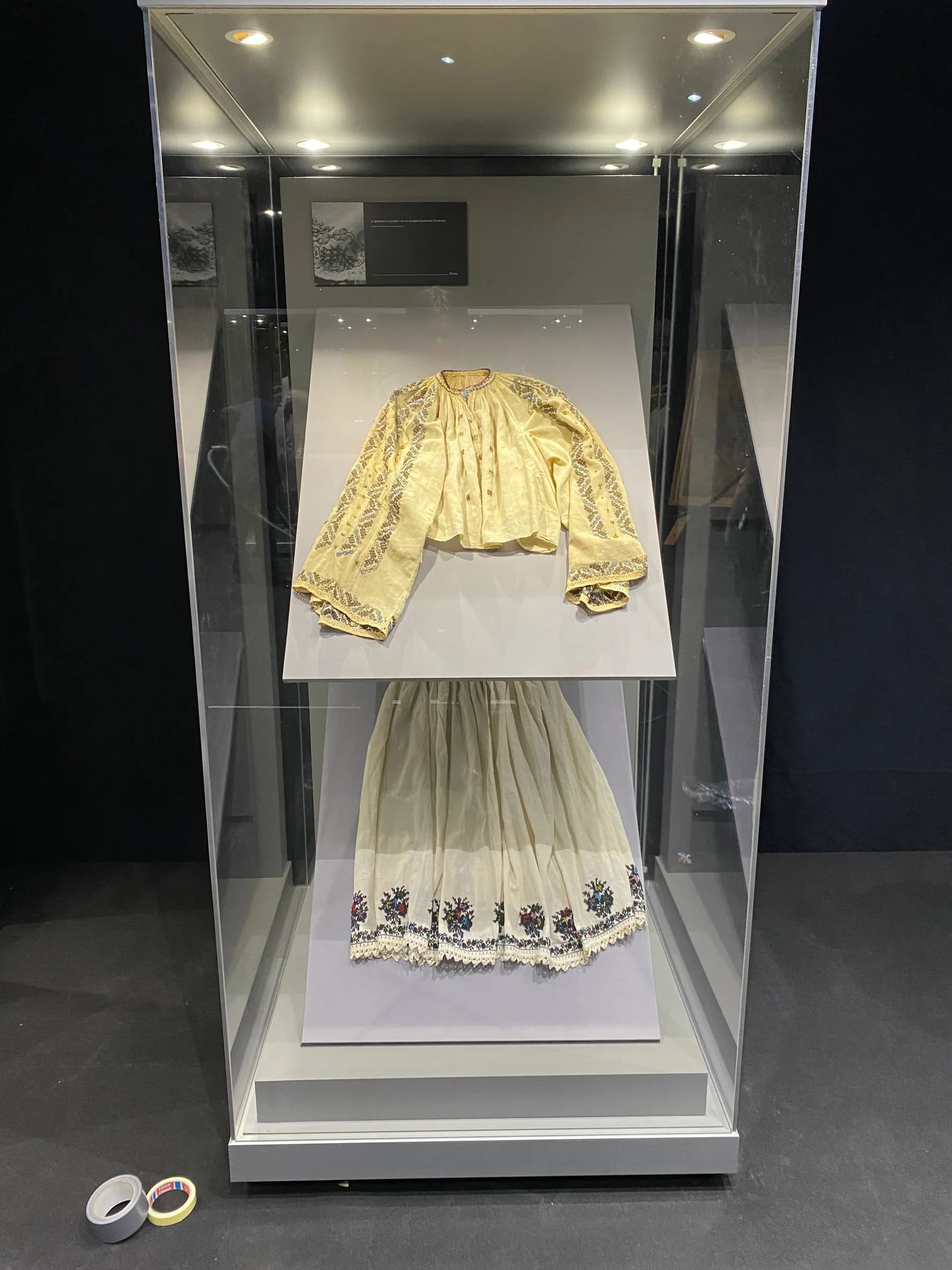 Tall display case with an embroidered blouse and skirt, lit from the top of the case