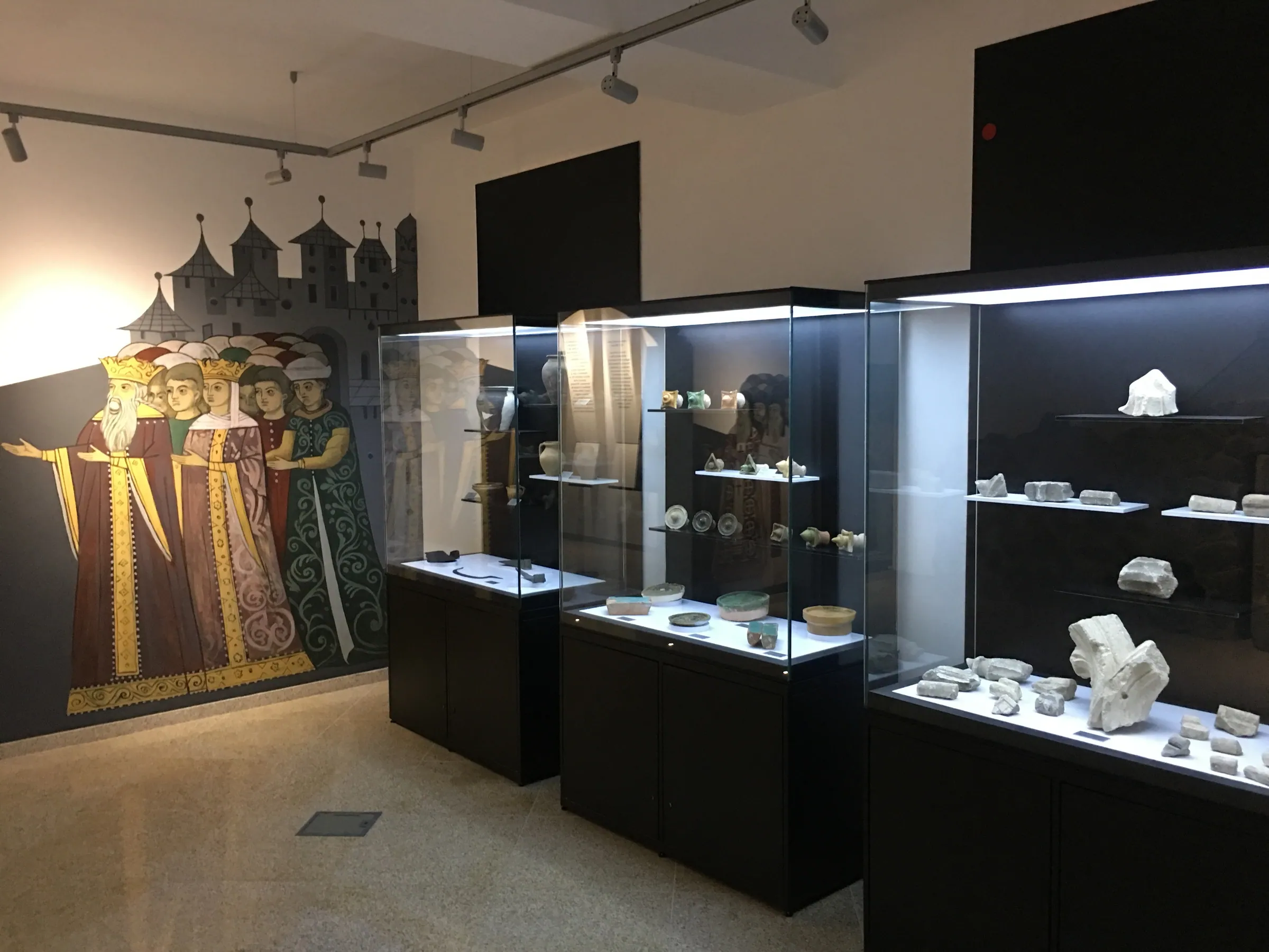 Display cases with stone fragments and objects, next to a mural of medieval figures