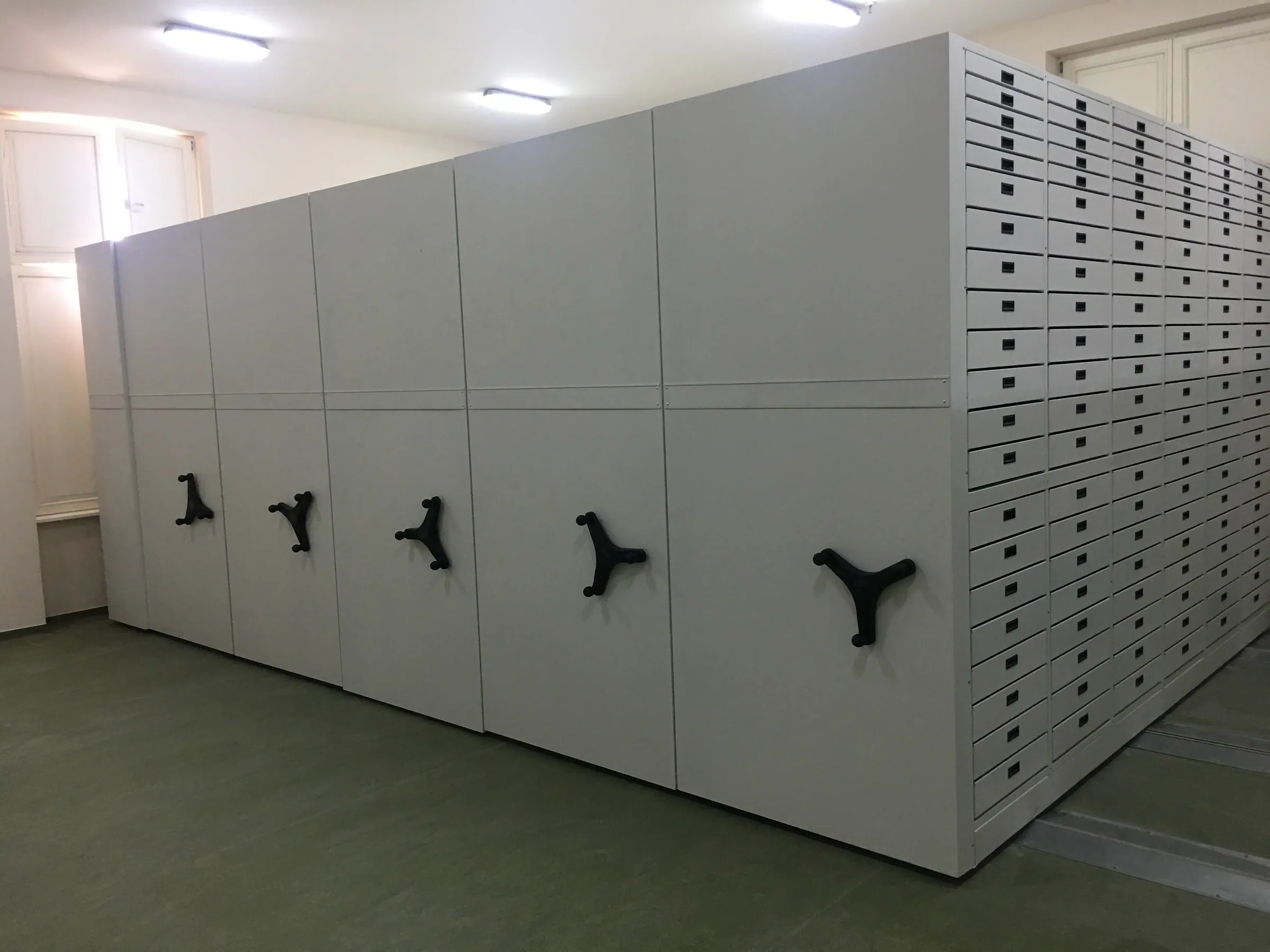 Grey mobile compact shelving with handwheels and flat drawers on the side